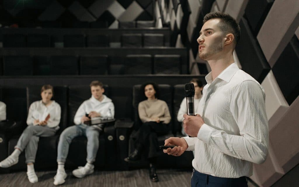 Ein Schauspieler mit einem Mikrofon steht vor einer Gruppe sitzender Personen