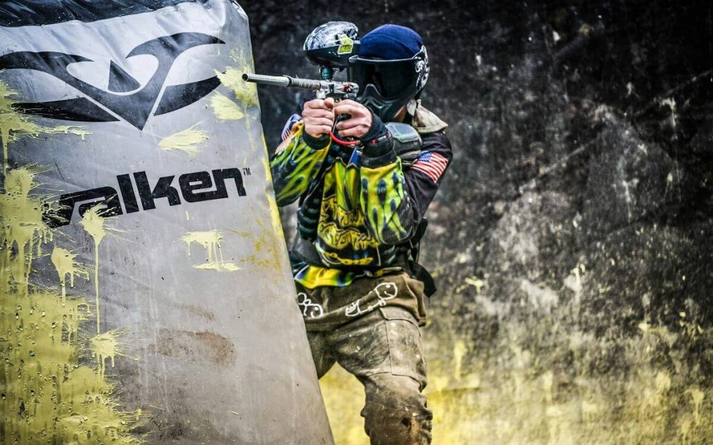 Eine Person mit Helm und Paintball-Pistole hinter einem mit Farbe besprühten Hindernis